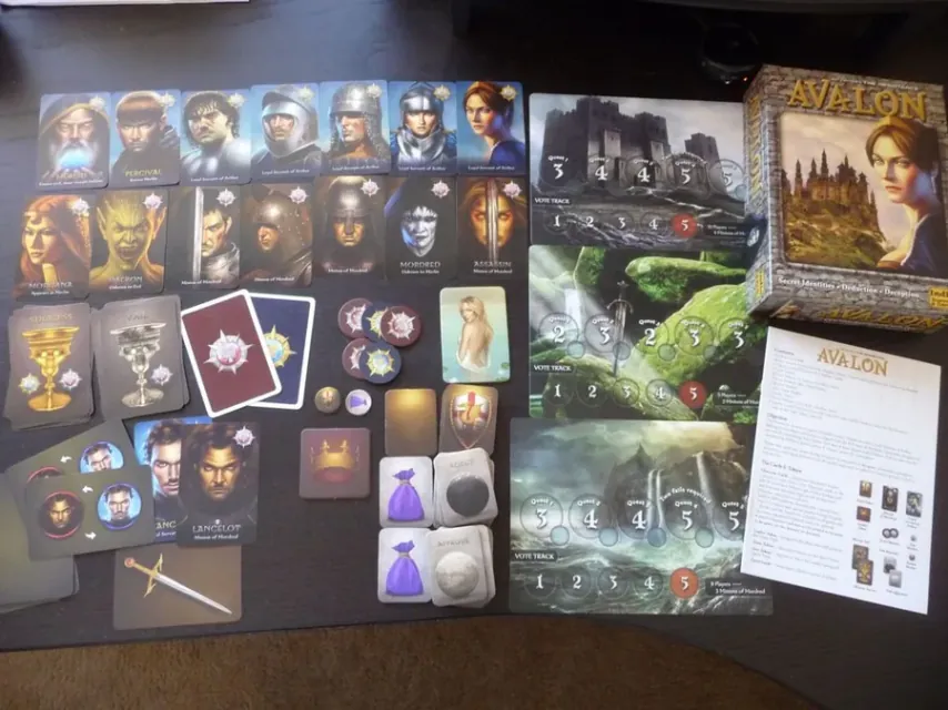 Todos los componentes de Avalon extendidos sobre la mesa: cartas de personaje, tablero, fichas de misión, de equipo y de voto, y marcadores.