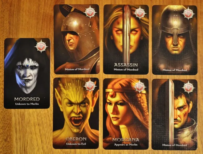 Cartas de personaje malvadas: Mordred, el Asesino, Oberon, Morgana y dos Esbirros de Mordred sobre una mesa de madera.