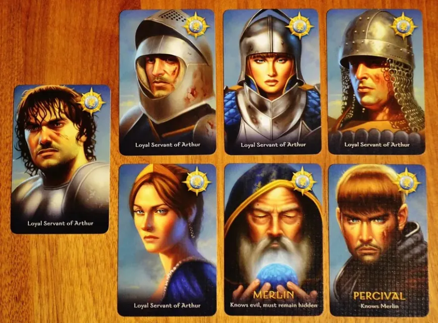 Cartas de personaje buenas: cinco Servidores Leales de Arturo, Merlín y Percival sobre una mesa de madera.